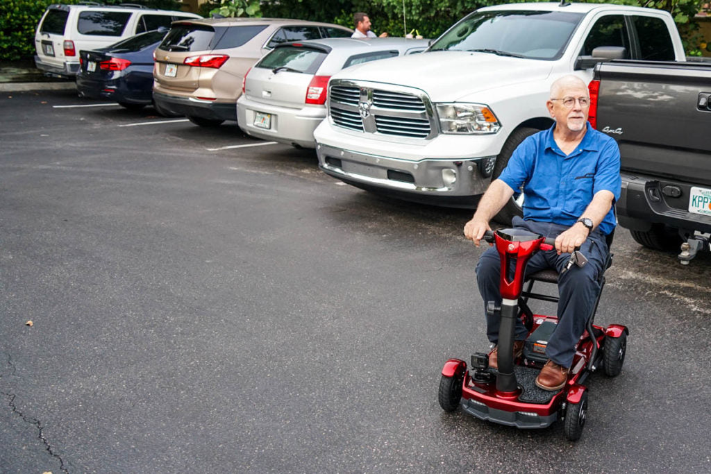 Man using a red EV Rider TEQNO folding mobility scooter in a parking lot with cars around