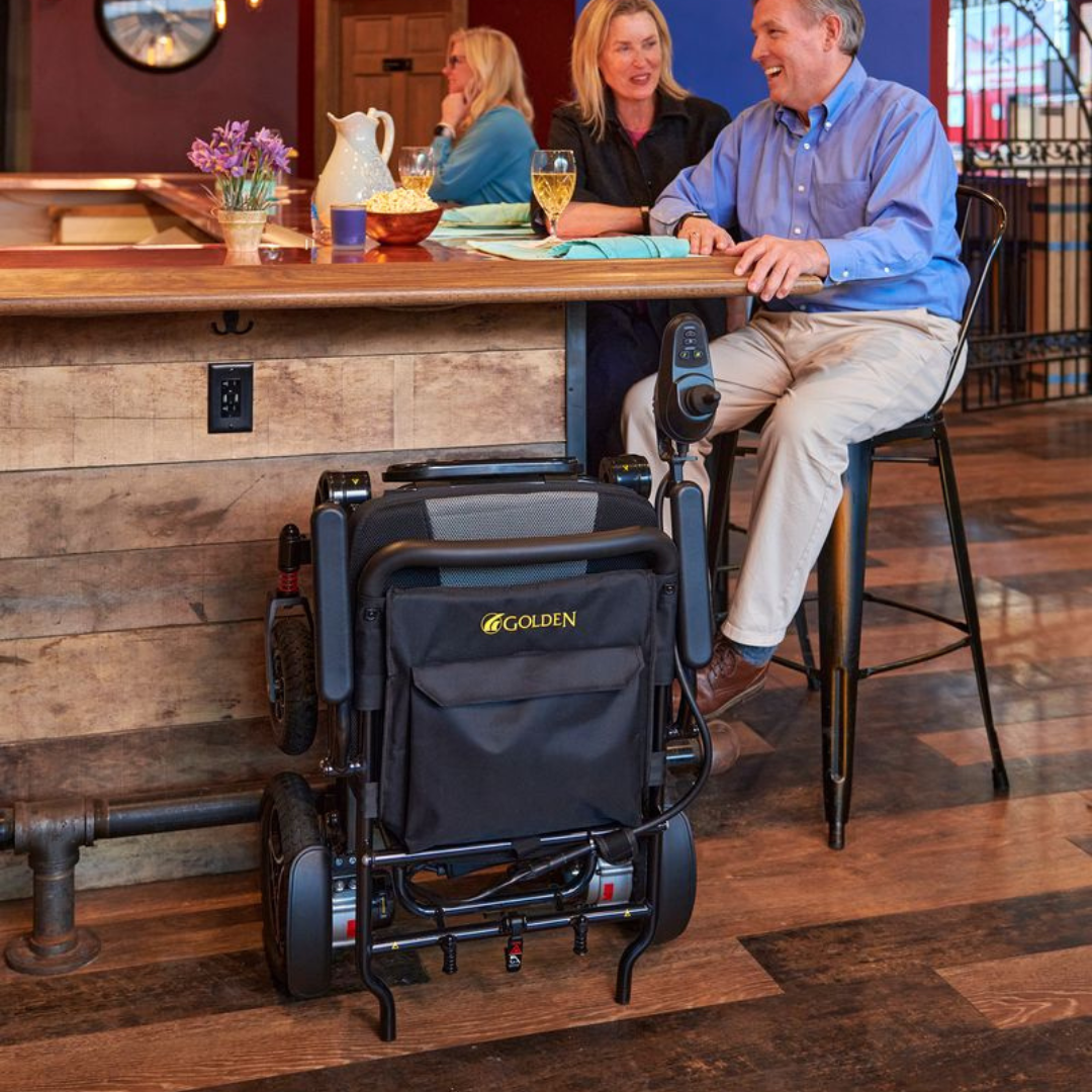 Person using a Golden Technologies Stride Folding Lightweight Electric Wheelchairat a bar with a wooden counter and patrons in the background.