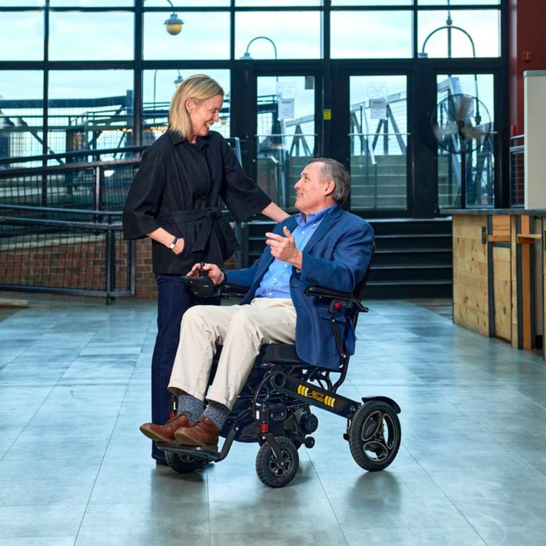 Man in a Golden Technologies Stride Folding Lightweight Electric Wheelchair being assisted by a woman in a modern indoor setting