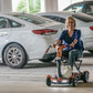Woman using a EV Rider TEQNO folding mobility scooter in gold in a parking garage with cars around