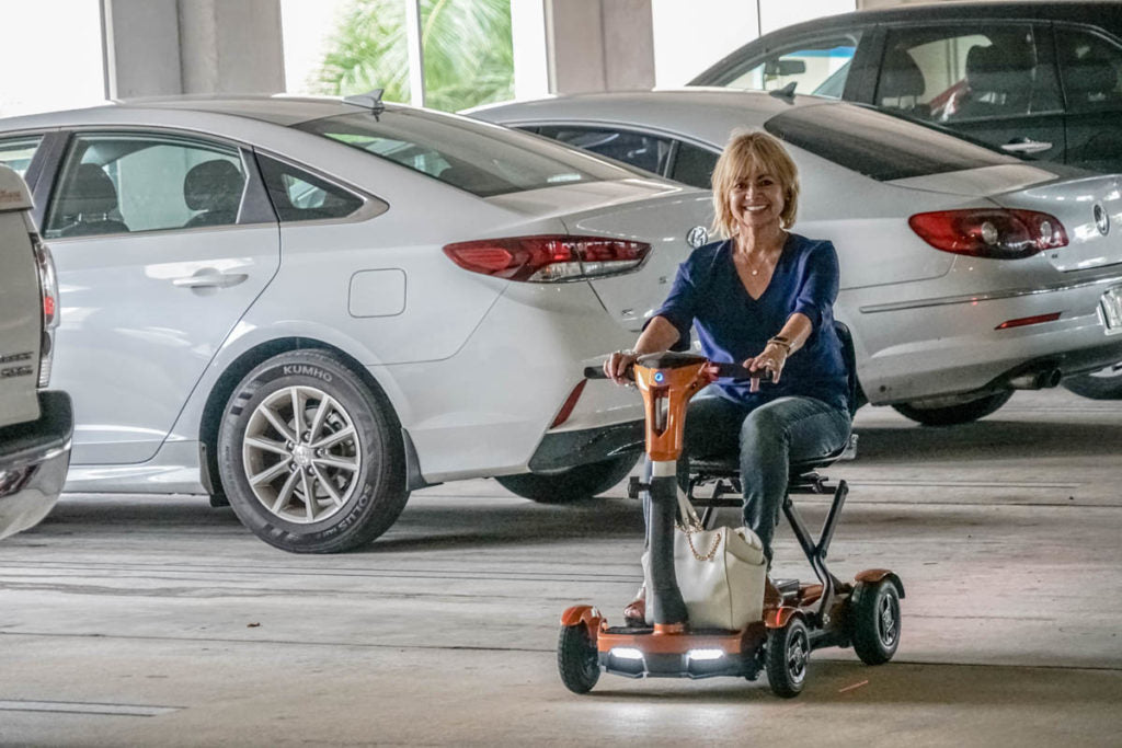 Woman using a EV Rider TEQNO folding mobility scooter in gold in a parking garage with cars around