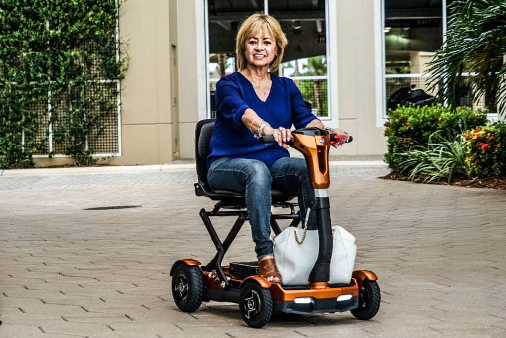 Woman using a EV Rider TEQNO folding mobility scooter in gold outdoors on a paved area.