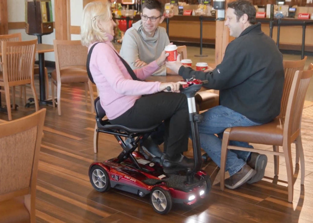 Woman sitting on EV Rider Transport 4AF Automatic Folding Travel Mobility Scooter in Red at lunch with friends.