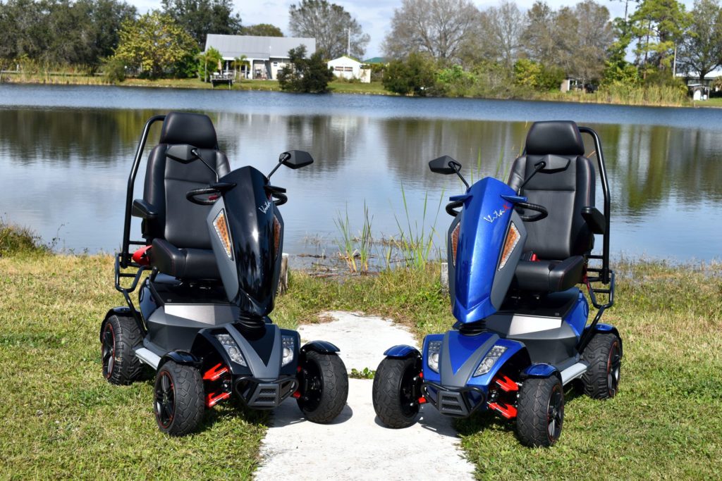 EV Rider Vita Monster Heavy-Duty Mobility Scooters on a grassy area near a body of water with trees in the background.
